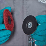 Two gloved hands holding circular abrasive Roloc-type discs against a gray work jacket background. One disc is red with a black center, and the other is black with a silver metal center.
